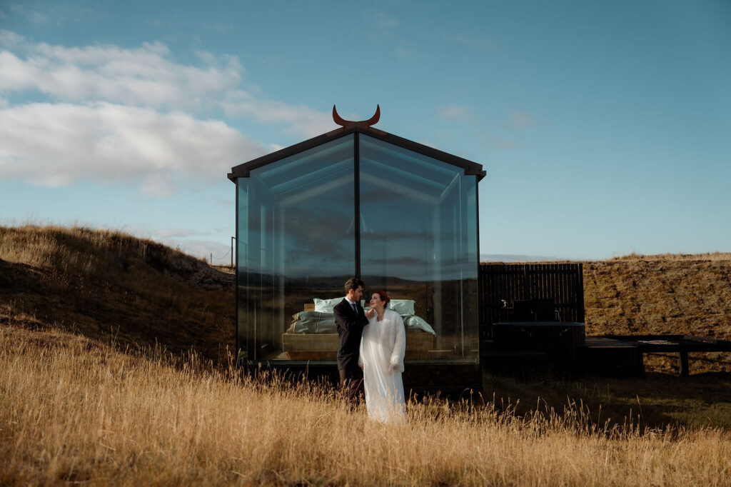bride and groom stand in front of a glass cabin in a grassy field, sharing a tender moment in iceland
