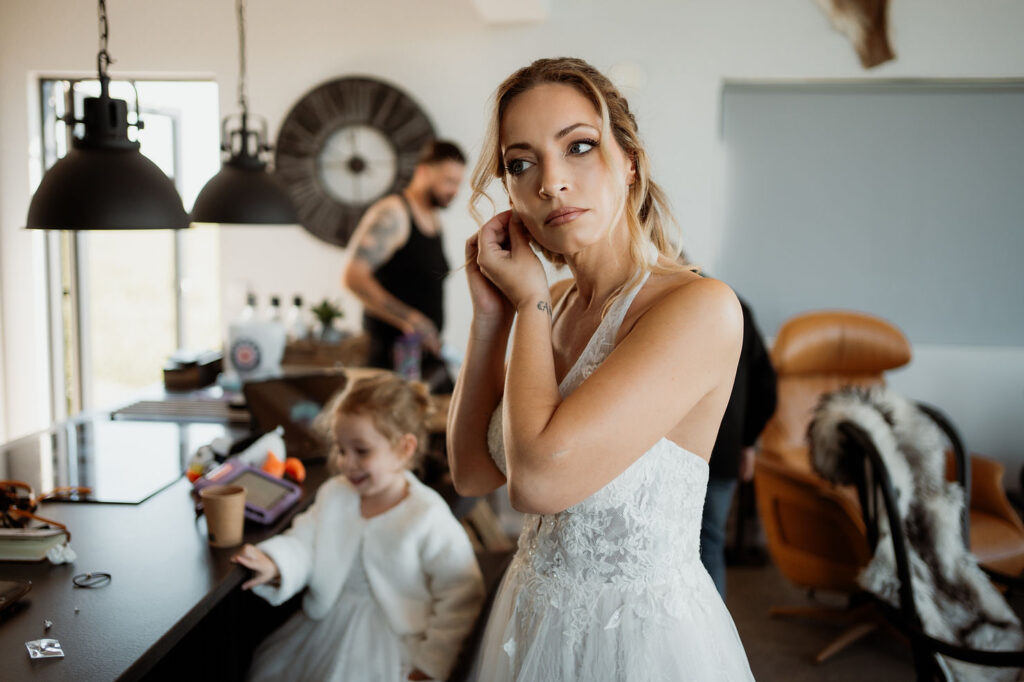 Bride in lace dress adjusts earring in cozy room with large clock. Child in white coat smiles during iceland elopement with family
