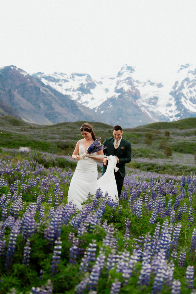Bride and groom in a field of purple flowers with snow-covered mountains in the background. Bride holding bouquet, groom holds train during iceland elopement