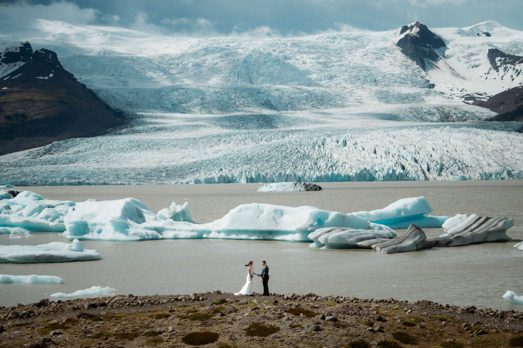 Bride and groom stand on rocky shore with icy lagoon and glaciers in the background during iceland wedding with family.