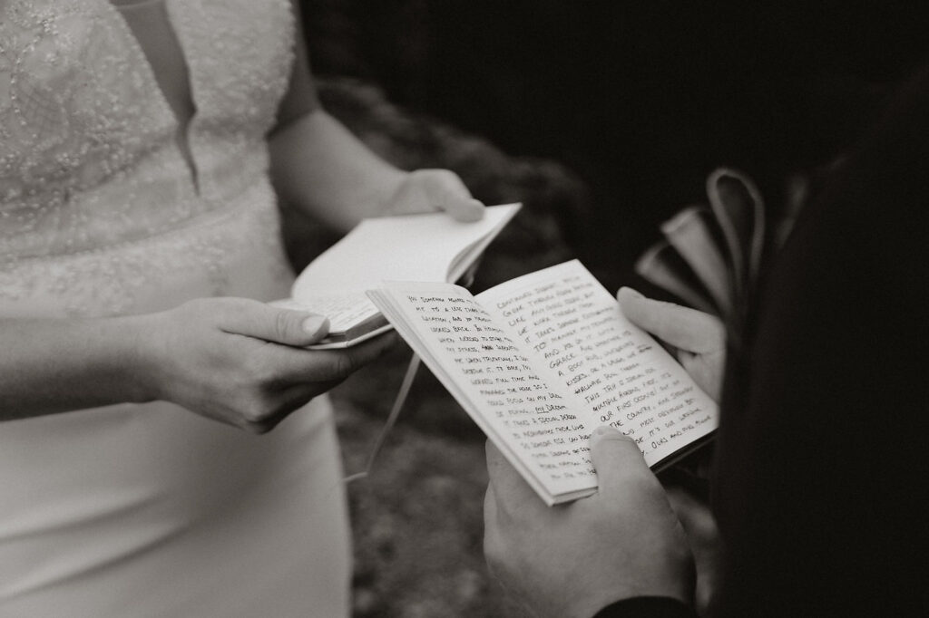 Close-up of a bride and groom exchanging handwritten vows in notebooks, wearing formal attire during iceland elopement with family included.