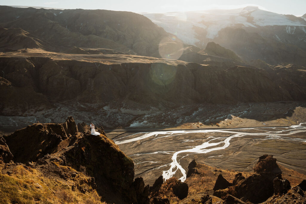 Thórsmörk: The Valley of Thor | How To Plan An Elopement Here: Bride and groom stand on a cliff in dramatic landscape, surrounded by rugged mountains and winding river.