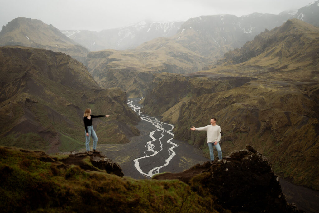 Thórsmörk: The Valley of Thor | How To Plan An Elopement Here: Two people stand on opposite cliffs, arms outstretched toward each other over a winding river, in a mountainous landscape under a cloudy sky.