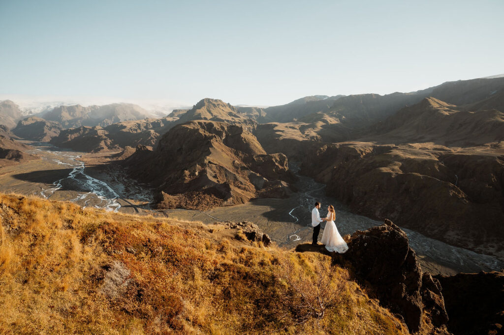 Þórsmörk Valley elopement: bride and groom stand on a cliff in wedding attire, holding hands against a vast mountainous landscape under a clear blue sky.