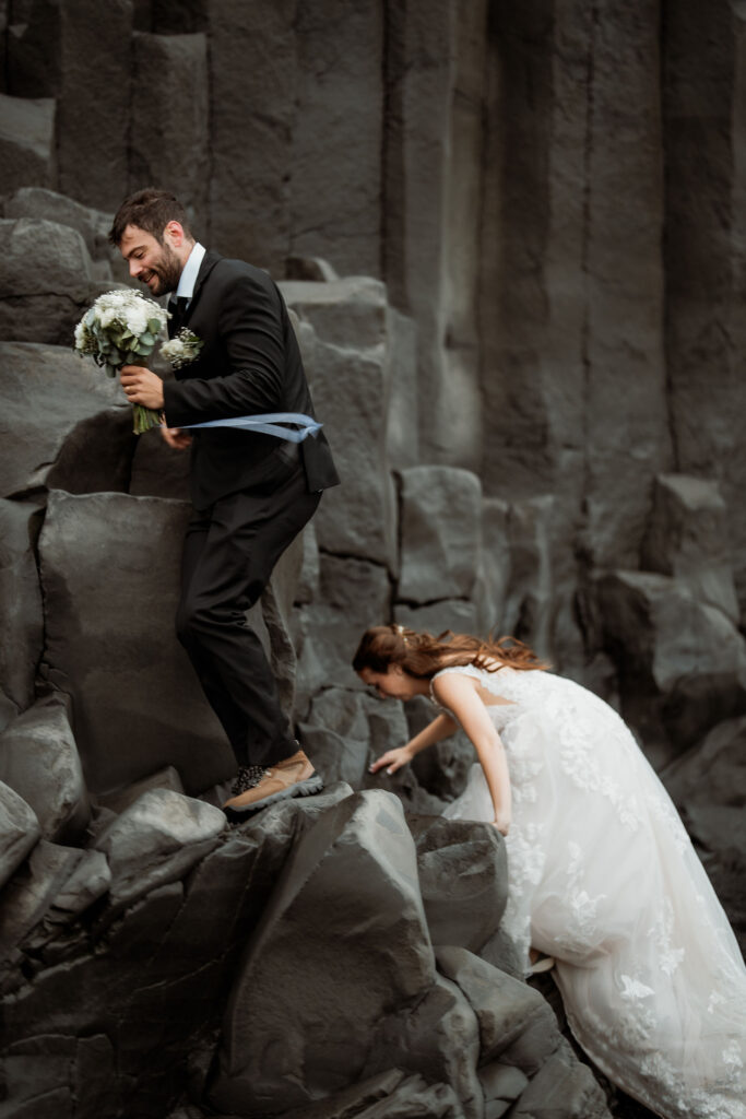 Þórsmörk Valley elopement: Bride and groom climb gray rocks; groom holds bouquet, bride in lace dress follows. Moody outdoor setting, showcasing adventure.