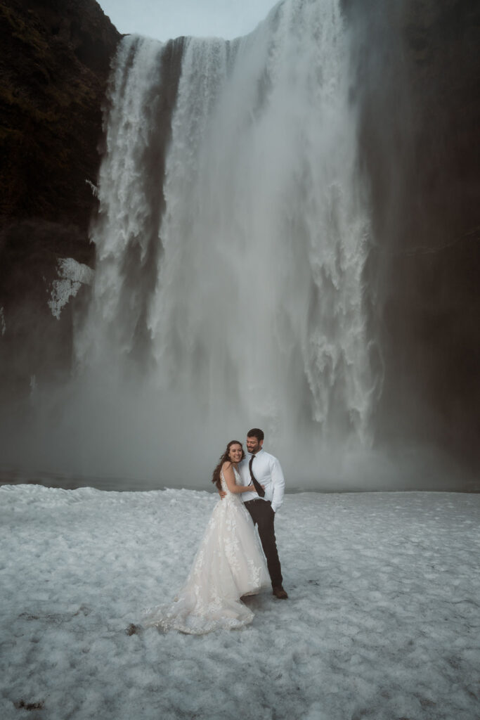 Bride and groom embrace on snowy ground in front of a massive waterfall at Þórsmörk Valley. The bride wears a flowing dress, and both smile joyfully.