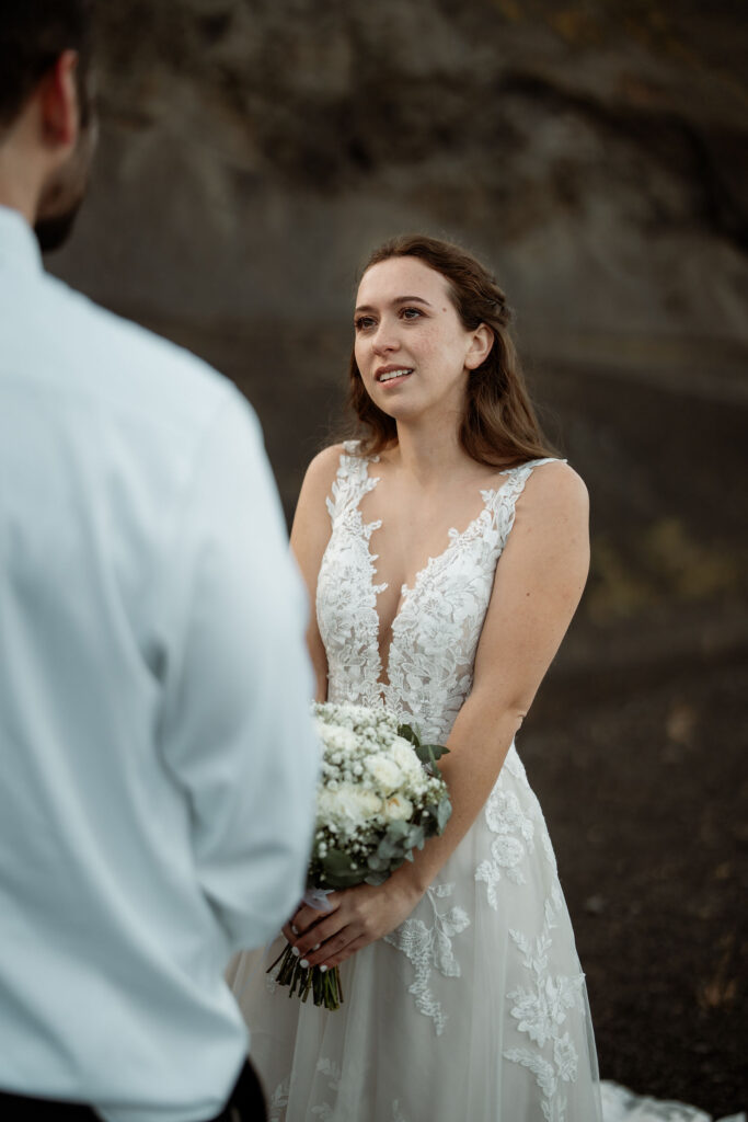Bride in a lace dress holds flowers, facing a groom in a white shirt. They stand on rocky terrain, exchanging vows at Þórsmörk Valley.