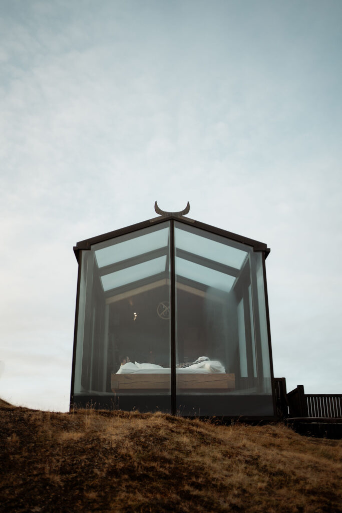 Glass cabin with a bed inside, set on a grassy hill under a cloudy sky at Þórsmörk Valley in iceland