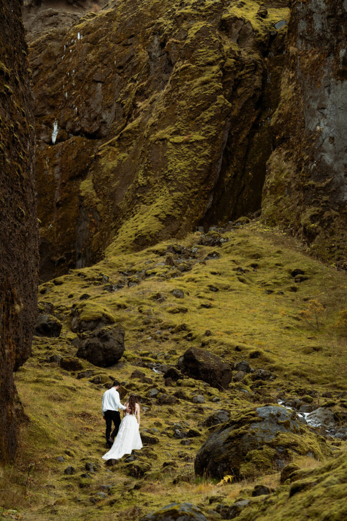 A bride in a white dress and a groom in a black suit walk hand in hand on a mossy, rocky landscape, surrounded by towering cliffs. Þórsmörk Valley