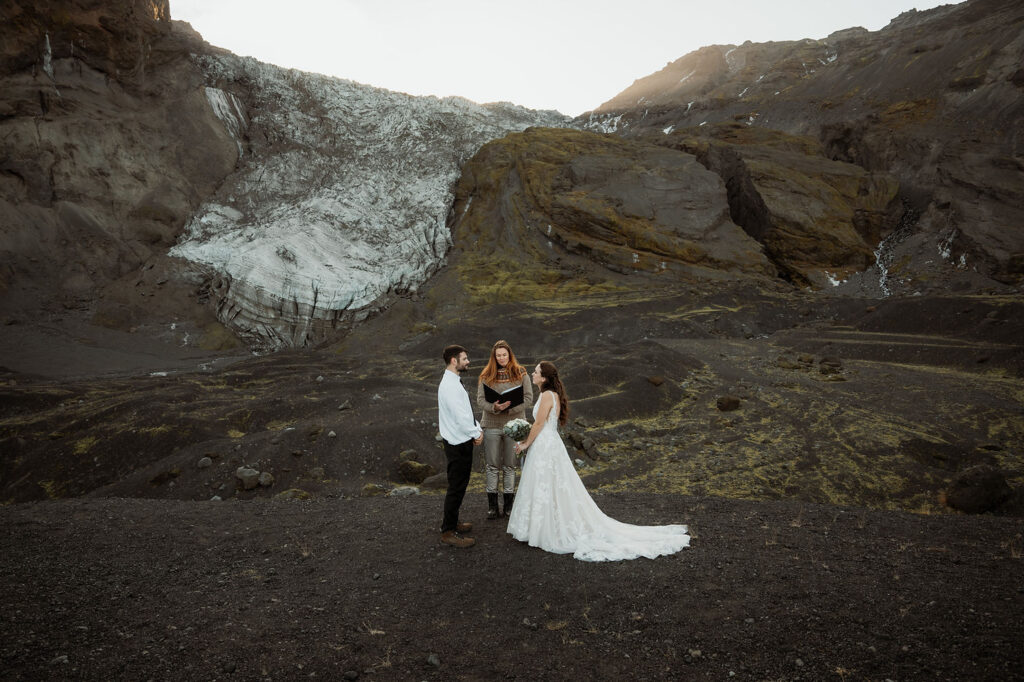 Bride and groom stand with officiant on rocky landscape with glacier backdrop. The bride wears a white gown, at Þórsmörk Valley in iceland.