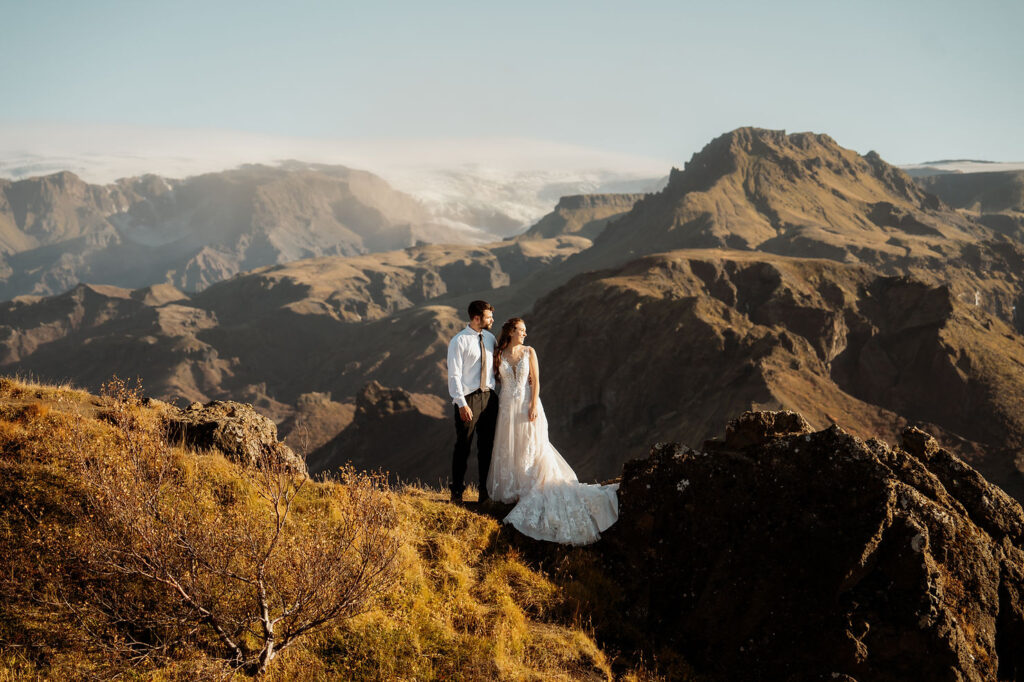 Bride and groom stand on a rocky cliff, surrounded by majestic mountains under a clear blue sky. Þórsmörk Valley in iceland.