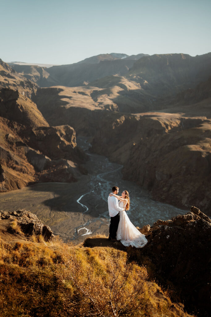 A couple embraces on a rocky cliff, overlooking a vast canyon with a meandering river. Þórsmörk Valley in iceland.