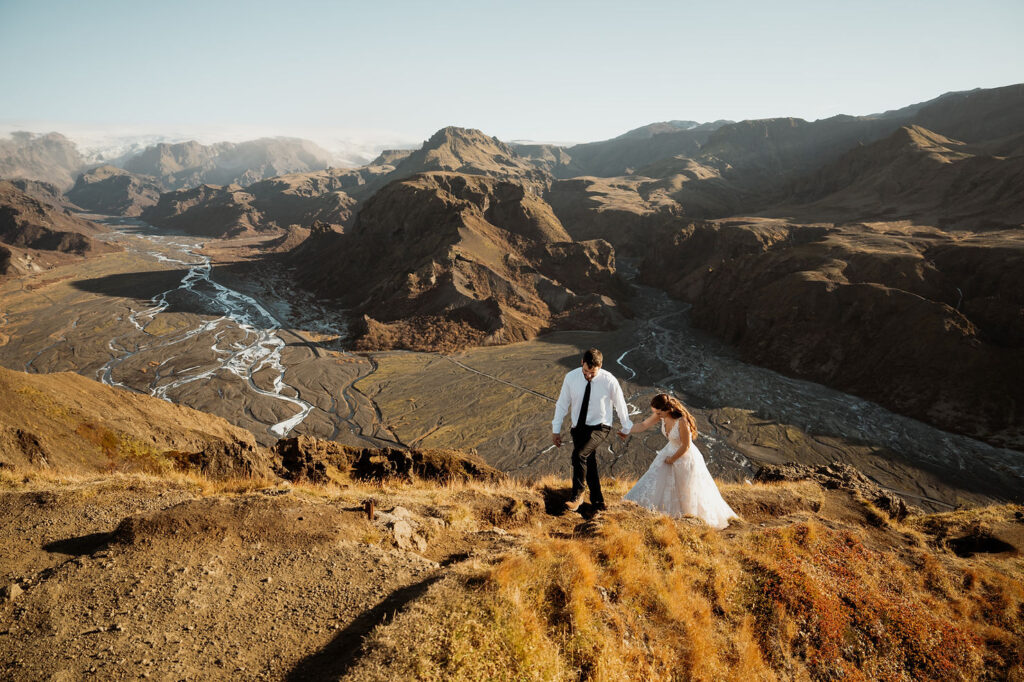 Bride and groom hold hands, ascending a grassy hill against a vast, mountainous landscape. Þórsmörk Valley in iceland.