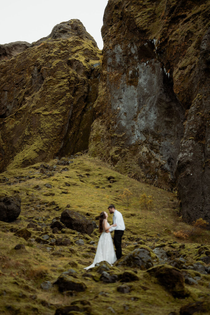 bride and groom embraces on a mossy hillside beneath rocky cliffs. Bride in a white dress and groom in black pants. Þórsmörk Valley in iceland