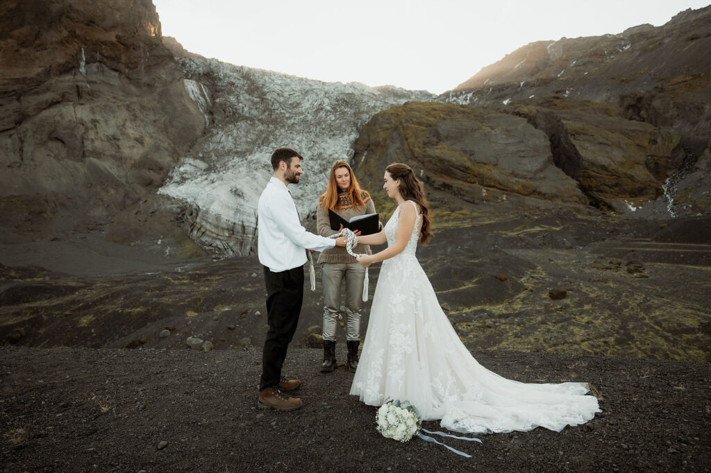 Bride and groom hold hands in a mountainous landscape, officiated by a woman holding a book. Þórsmörk Valley in iceland.
