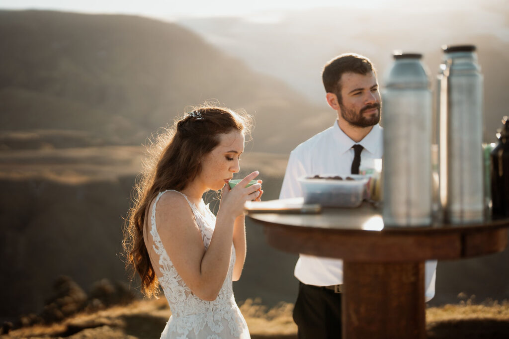 bride sips from a cup, standing by groom next to a table with thermoses. Sunlit mountains in the background at Þórsmörk Valley in iceland.