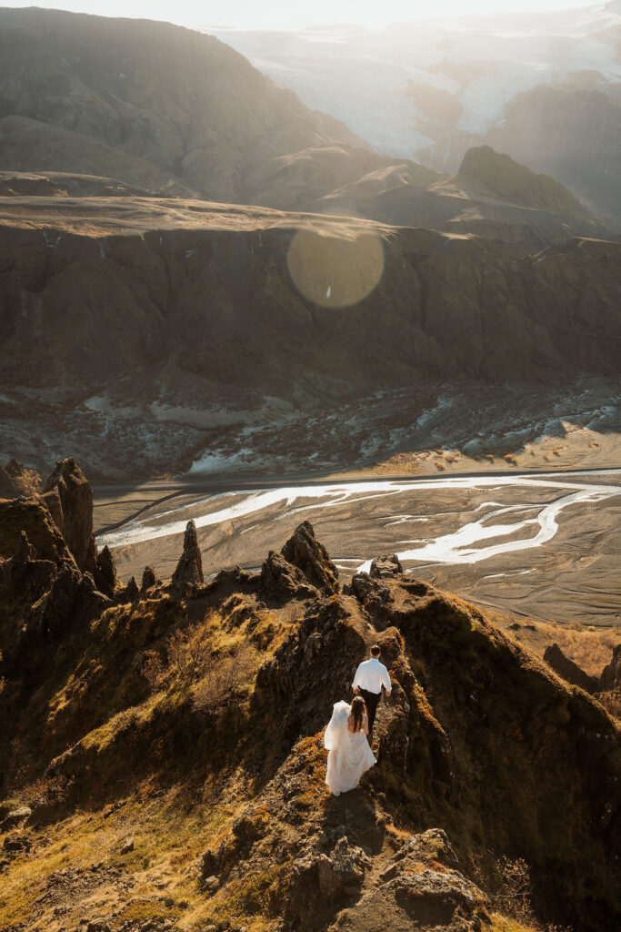 bride and groom walk along a Þórsmörk Valley with a river and mountains in the background, bathed in warm sunlight. The bride wears a white dress.
