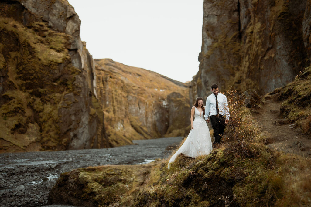 Bride and groom walk hand in hand on a grassy cliff, surrounded by rocky canyon walls. Þórsmörk Valley in iceland.