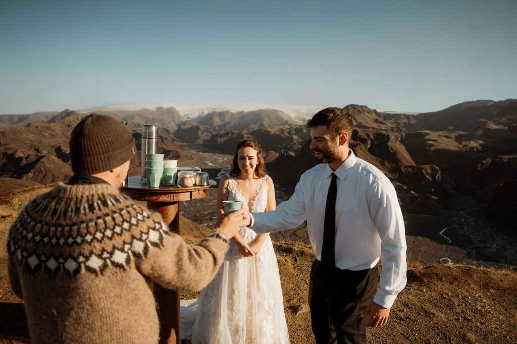 Bride and groom smile while sharing elopement drinks with a man in a knit sweater. Mountainous landscape of Þórsmörk Valley, iceland in the background.