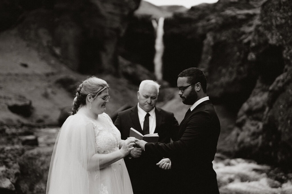 How to Include Your Family When You Elope: Bride and groom exchange rings in a serene outdoor setting with a waterfall in the background. An officiant holds an open book. Black and white.