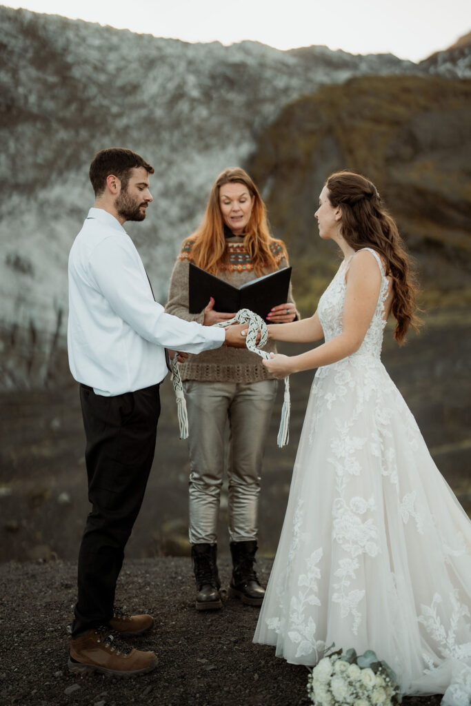 Bride and groom hold hands with a cord during a ceremony in a mountainous landscape. An officiant reads from a book. Mood is serene.
