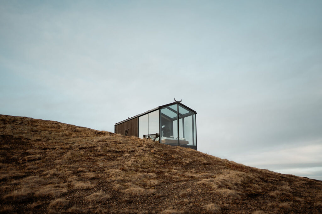 Glass cabin with wooden accents on a grassy hill under cloudy blue sky; for a Þórsmörk Valley elopement in iceland.