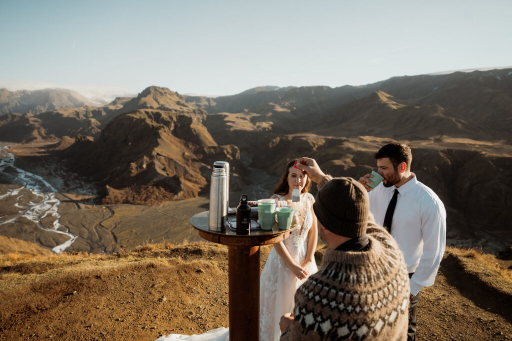 bride and groom drink tea on a cliff with rugged mountains in the background. A man holds a teabag. at Þórsmörk Valley during iceland elopement
