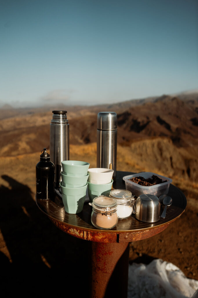 Thermoses, cups, jars, and a food container on a rustic table set against a mountainous landscape of Þórsmörk Valley in iceland for wedding.