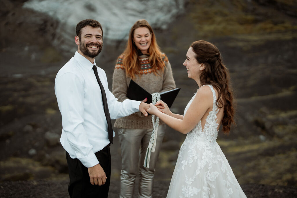 bride and groom exchange elopement vows at Þórsmörk Valley; the man wears a white shirt, the woman in a lace dress, an officiant behind holds a tied cord, smiling.