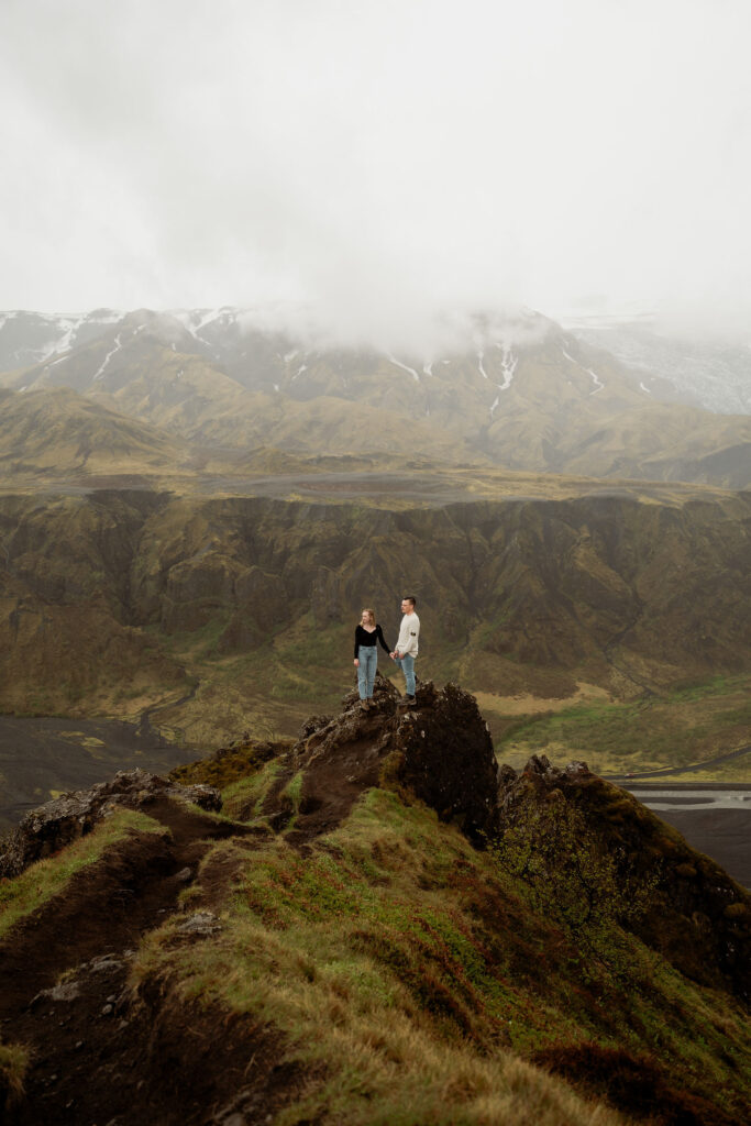Couple holding hands on a rocky cliff with misty mountains in the background. Overcast sky, earthy tones, and a serene mood at Þórsmörk Valley