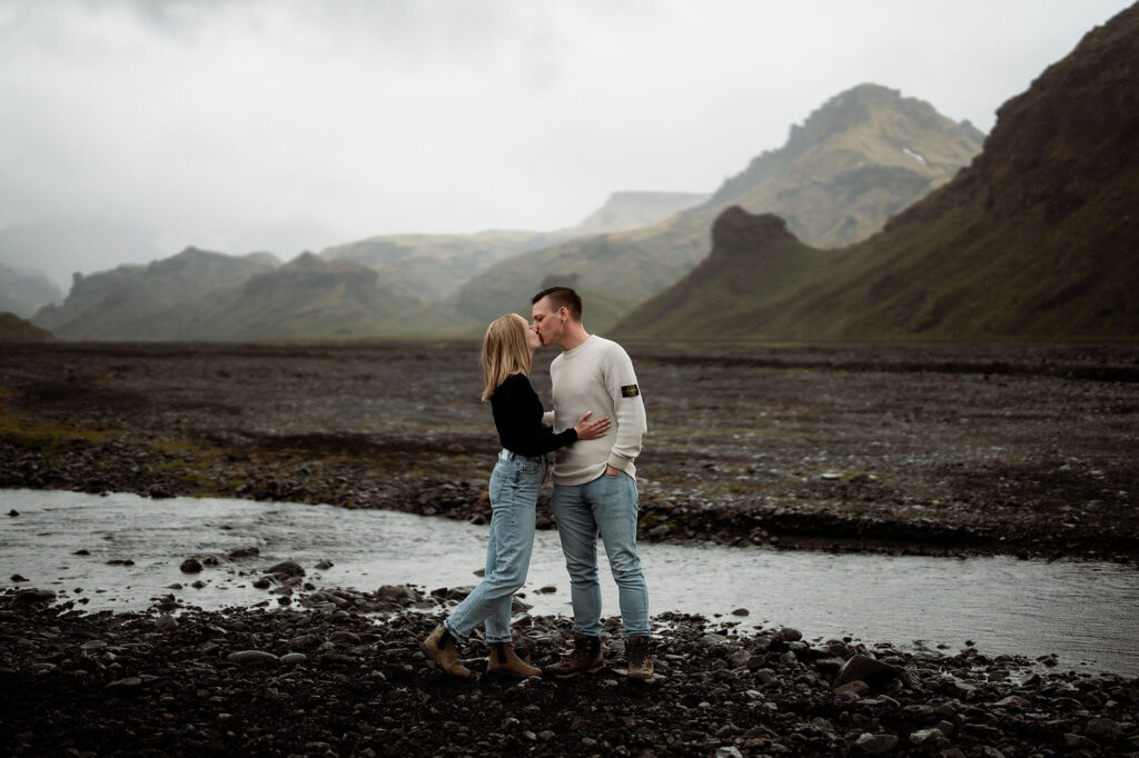 Couple kissing by a rocky stream with misty mountains in the background. Overcast sky, both wearing casual sweaters and jeans during engagement photos at Þórsmörk in iceland