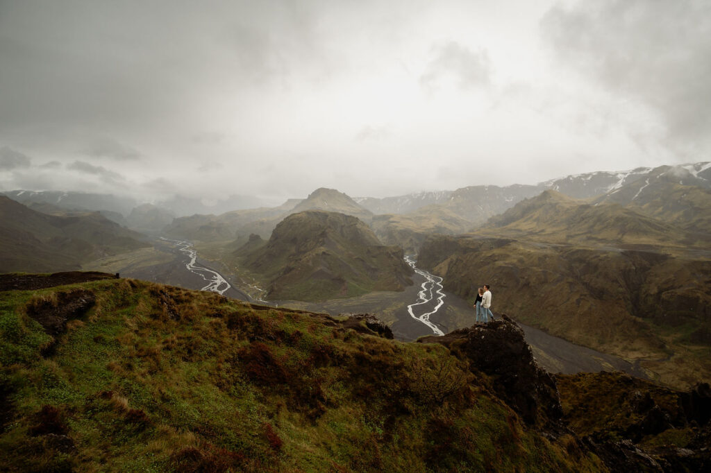 A couple stands on a grassy mountain cliff overlooking the Þórsmörk Valley with a winding river. 