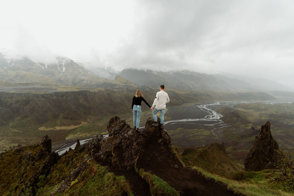 Couple holding hands on a rocky cliff, overlooking a vast, misty landscape with mountains and a winding river below.