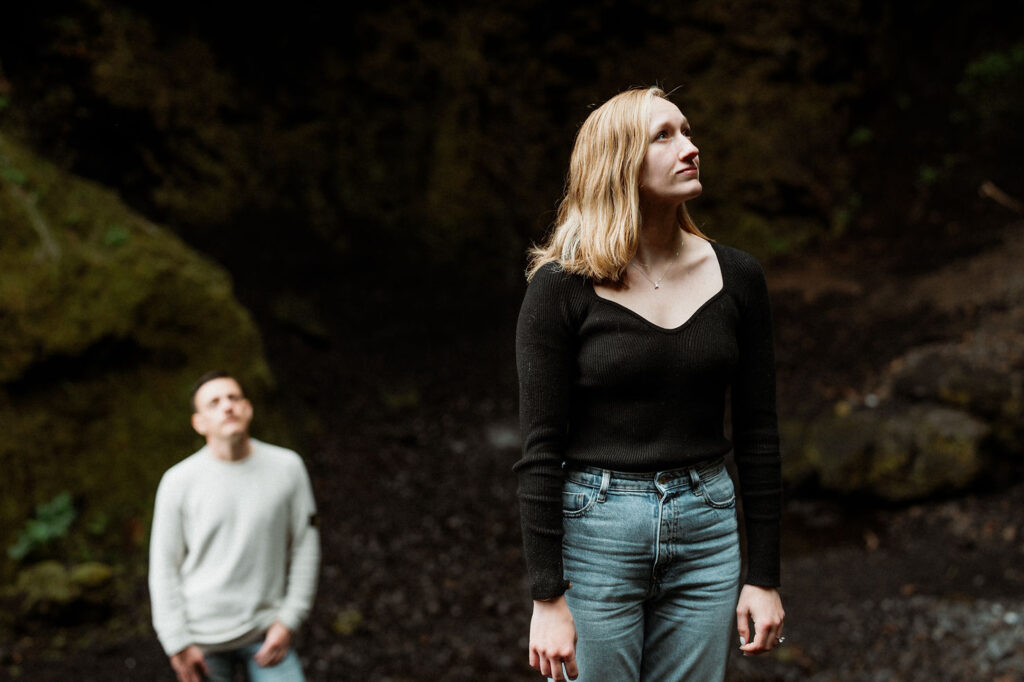 A woman in a black top stands in a forest looking upward. A man in a white sweater is blurred in the background during Þórsmörk Valley engagement photos