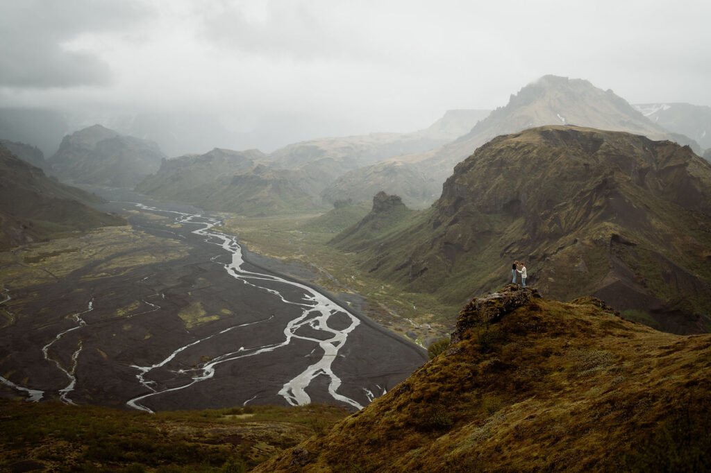 A couple stands on a grassy cliff in a vast, misty mountain landscape with winding rivers below, in the Þórsmörk Valley in iceland.