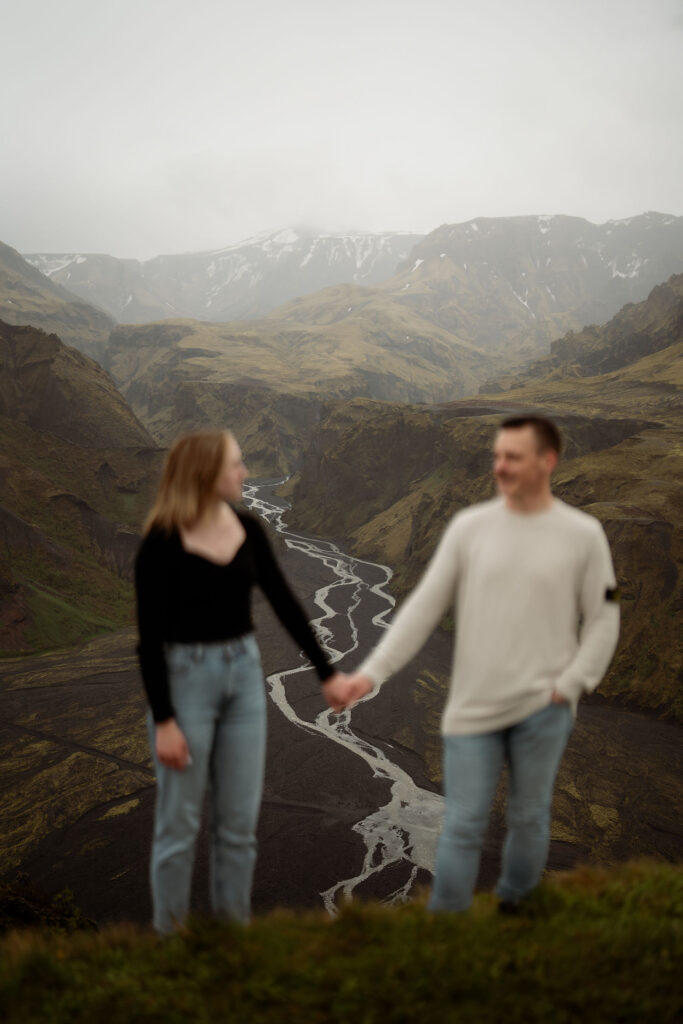 An engaged couple holds hands in the foreground, overlooking a foggy landscape with a winding river and grassy mountains in the Þórsmörk Valley