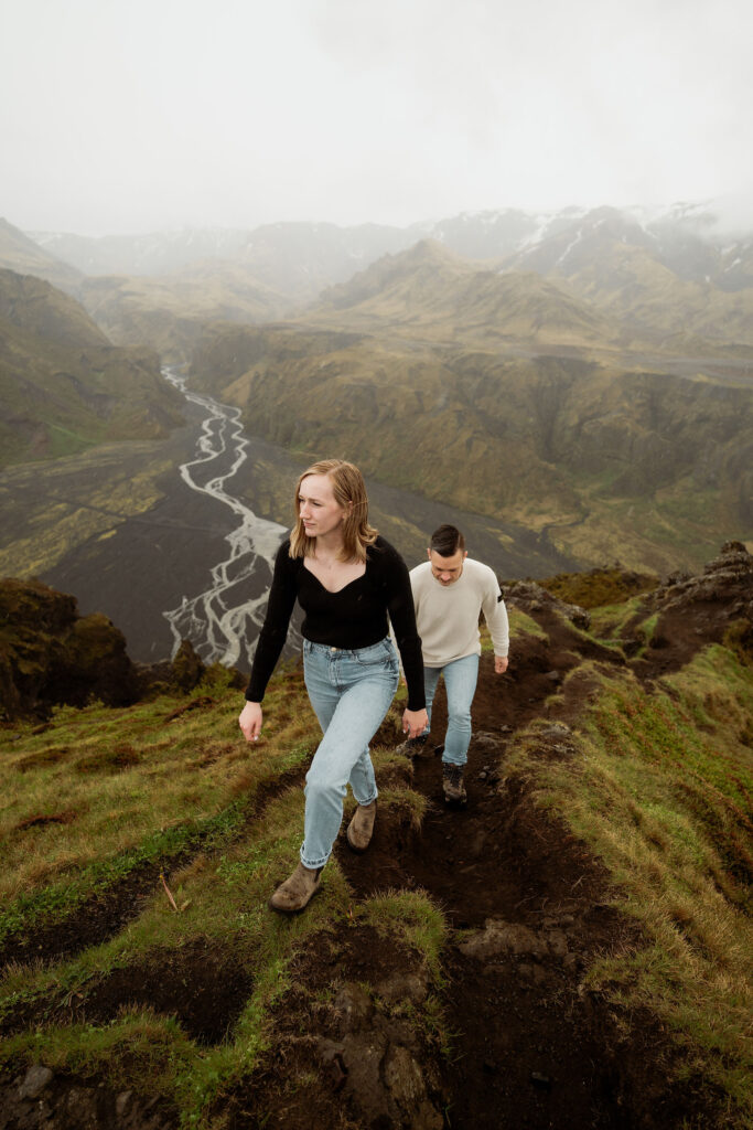engaged couple hike up a grassy mountain trail with a river and misty mountains in the background, at the Þórsmörk Valley