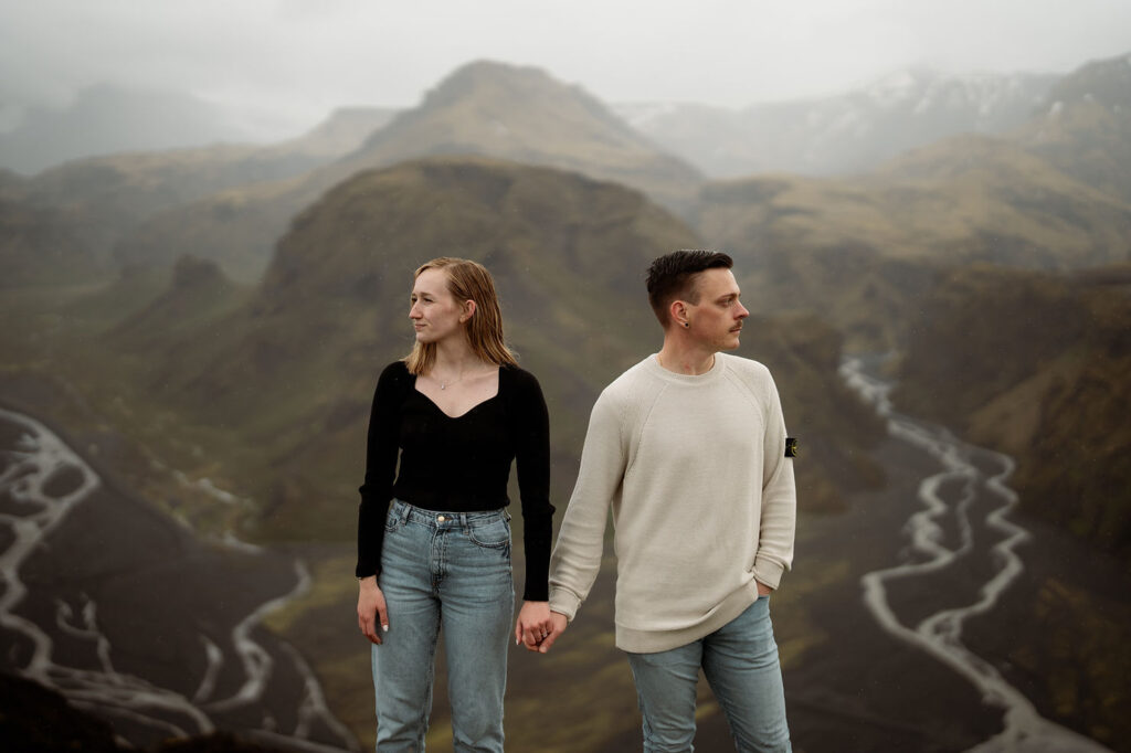 engaged couple holds hands, gazing opposite directions in front of the Þórsmörk Valley in iceland. River valleys in the background.