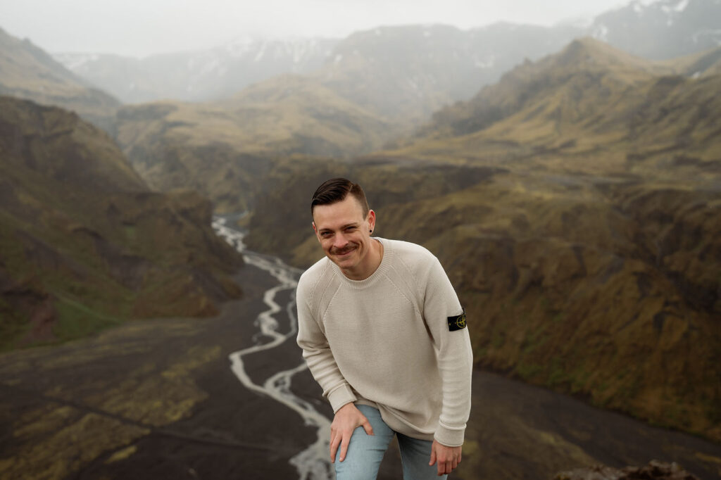 A person in a light sweater leans forward, smiling, with a scenic view of Þórsmörk Valley and a winding river in the background. 