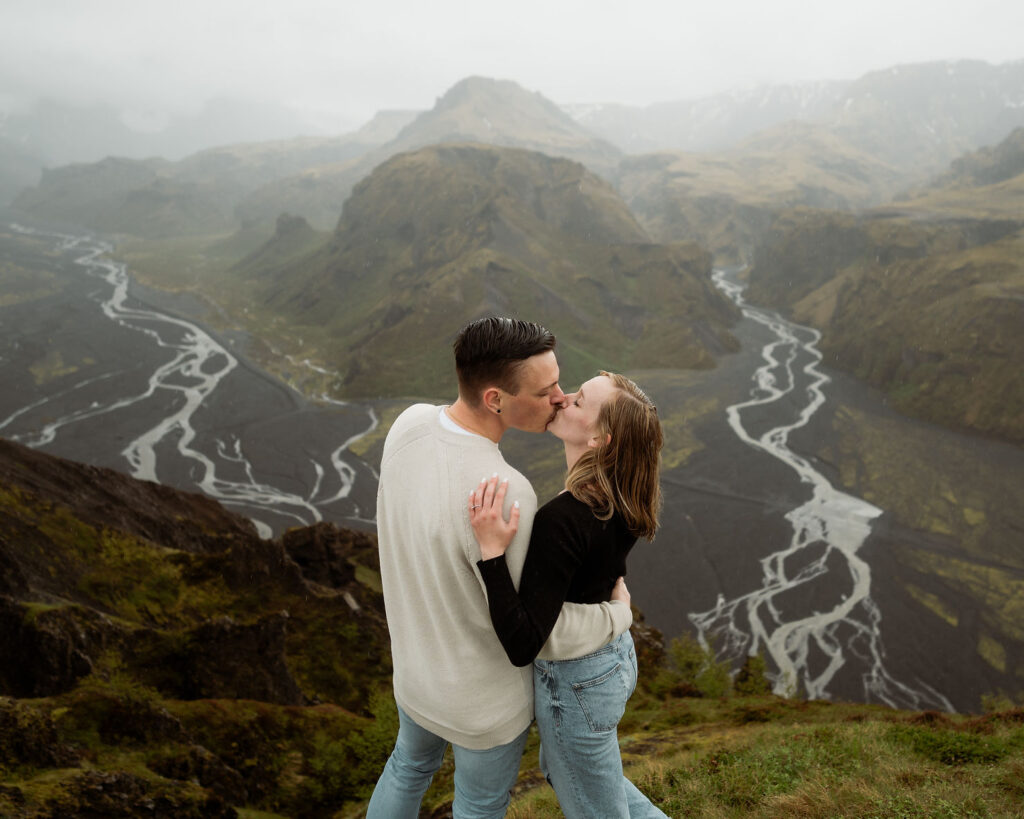 engaged couple kisses on a grassy hill overlooking the Þórsmörk Valley with winding streams and rugged mountains, under a cloudy sky in iceland.
