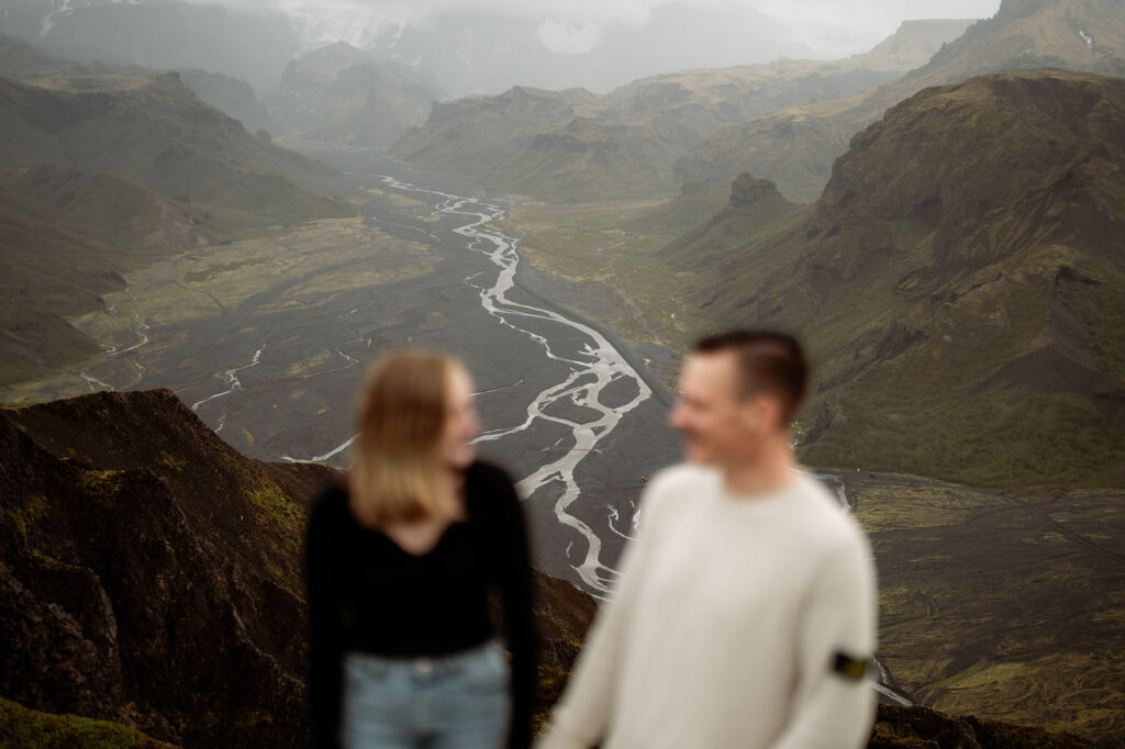 engaged couple smiling in the foreground with a blurred volcanic landscape and winding river in the background, under a cloudy sky in iceland