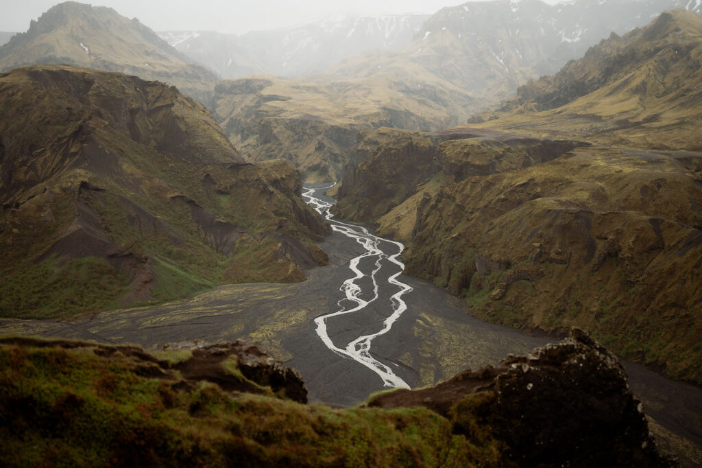 Winding river through rugged, mossy mountains with a cloudy sky in the Þórsmörk Valley. The mood is tranquil, featuring earthy browns and greens.