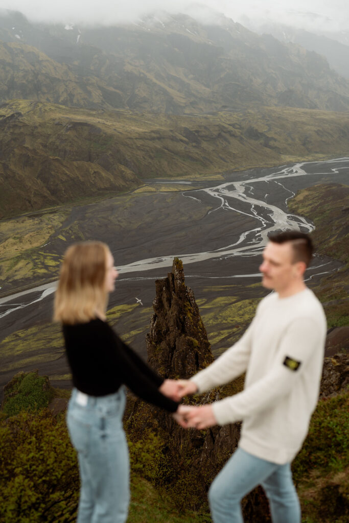 engaged couple holds hands in the foreground, blurred. They stand in front of the vast, misty landscape of Þórsmörk Valley in iceland