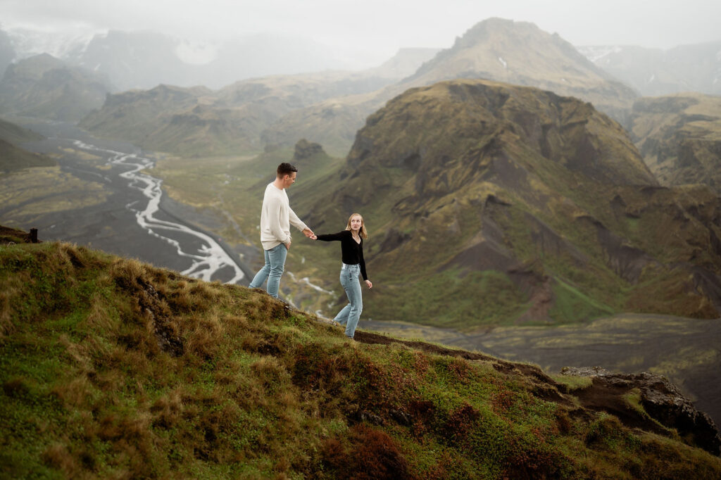 engaged couple holding hands walks on a grassy hill with a mountainous landscape and winding river in the background, creating a serene mood at Þórsmörk Valley