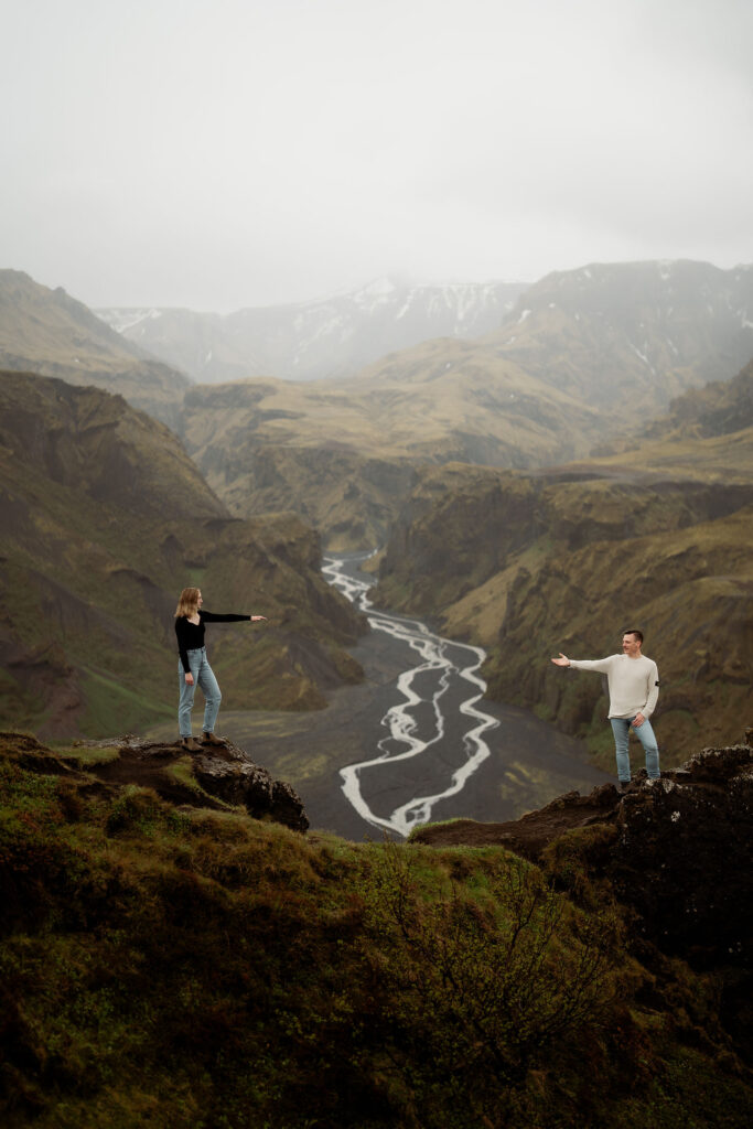 engaged couple stands on opposite cliffs in a misty valley, pointing across a winding river. Þórsmörk Valley during engagement photos.