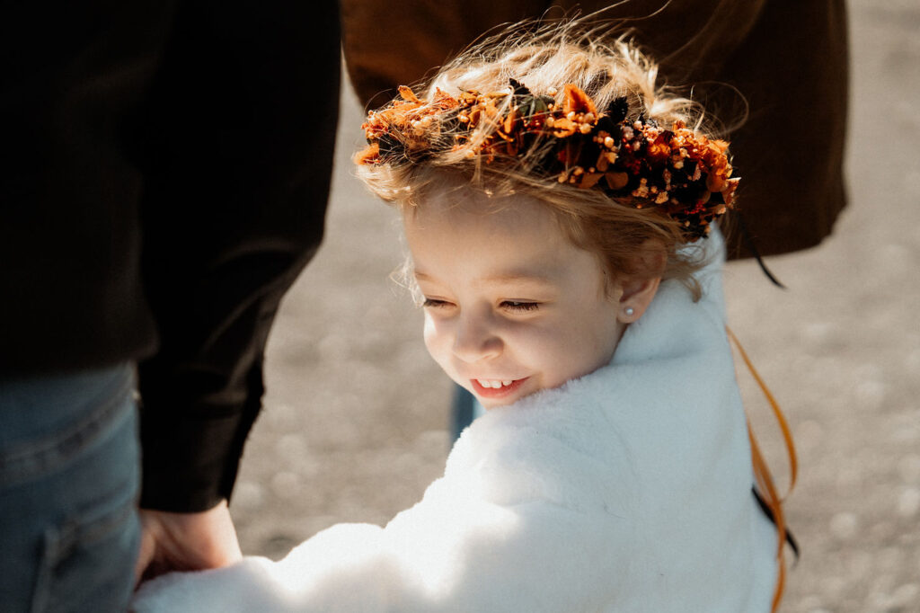 Young child in white coat smiling, wearing an orange floral crown, holding an adult's hand outdoors during an iceland elopement with family.