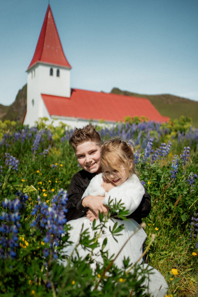 How to Include Your Family When You Elope: Two children smiling in a flower field, cuddling. In the background, a white church with a red roof peeks out under a clear blue sky.
