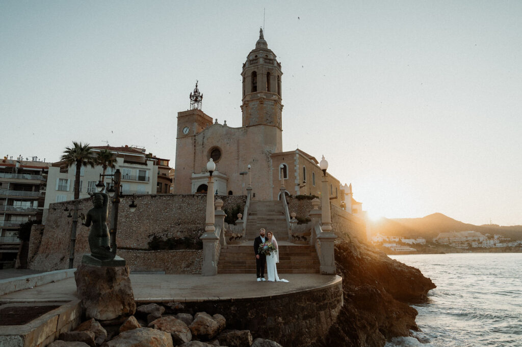 Bride and groom stand on a stone platform by a historic church in Barcelona at sunset, with ocean waves and a mermaid statue nearby. Peaceful setting.