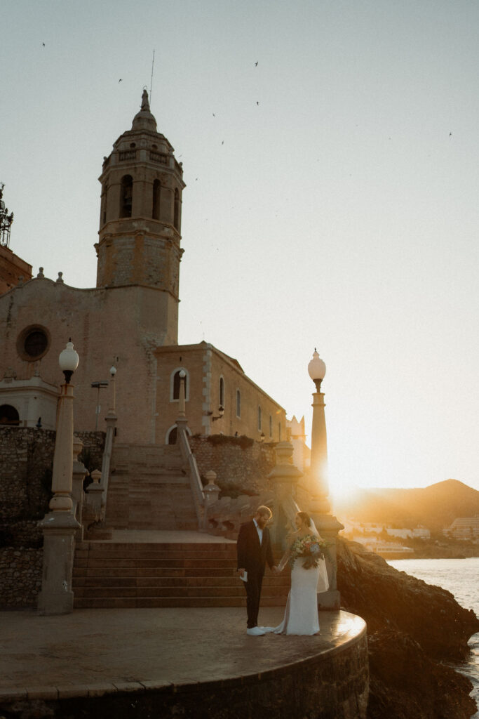 A couple holds hands at sunset by an old church during Barcelona elopement. The bride holds a bouquet, creating a romantic, serene scene.