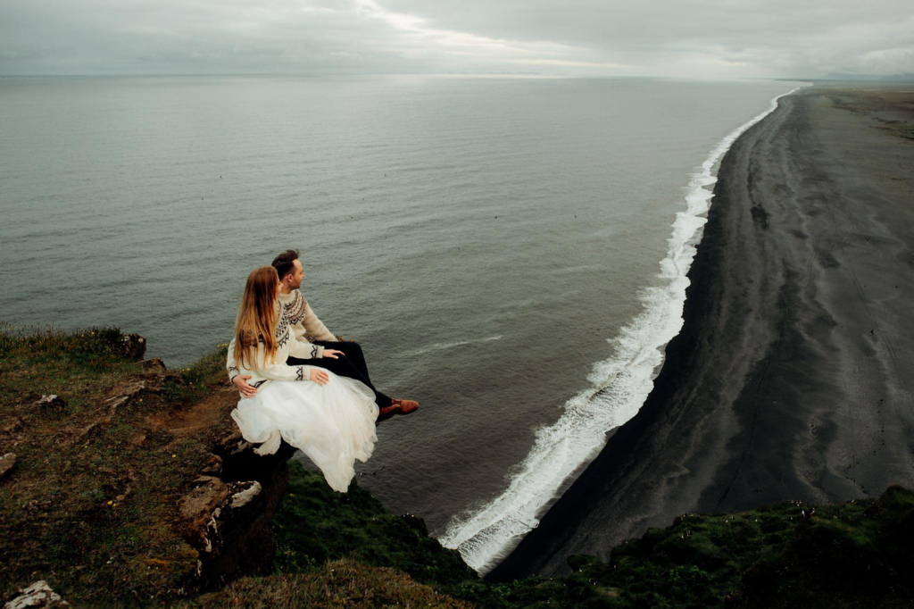 A couple sits on a cliff's edge overlooking a vast ocean and black sand beach. They're dressed warmly, creating a serene, romantic mood.